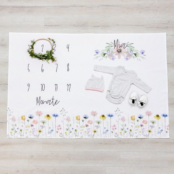 Meilensteindecke für das erste Lebensjahr mit buntem Blumenmuster, Monatskalender und personalisiertem Namen über diesem, hier mit Blätterkranz zur Markierung des Alters