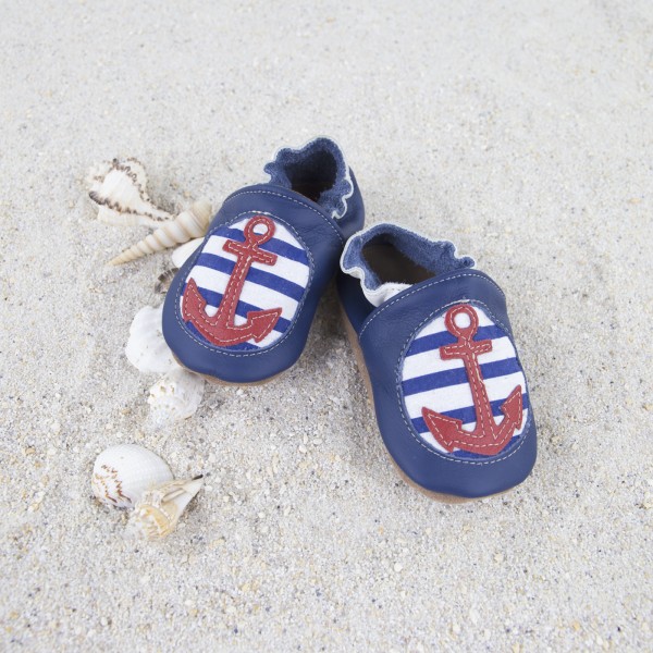 dunkelblaue Krabbelschuhe aus Leder mit rotem Anker auf blau-weiß gestreiftem Stoff vorne und hellbrauner Sohle, dekoriert auf Sand mit Muscheln